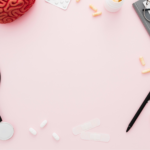 Medical-themed workspace on a pastel pink background, featuring a stethoscope, model brain, scattered pills, glasses, band-aids, a pen, and a notebook.